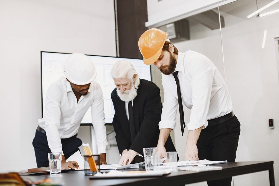 Group of architects discussing construction blueprints wearing hardhats in a modern office.
